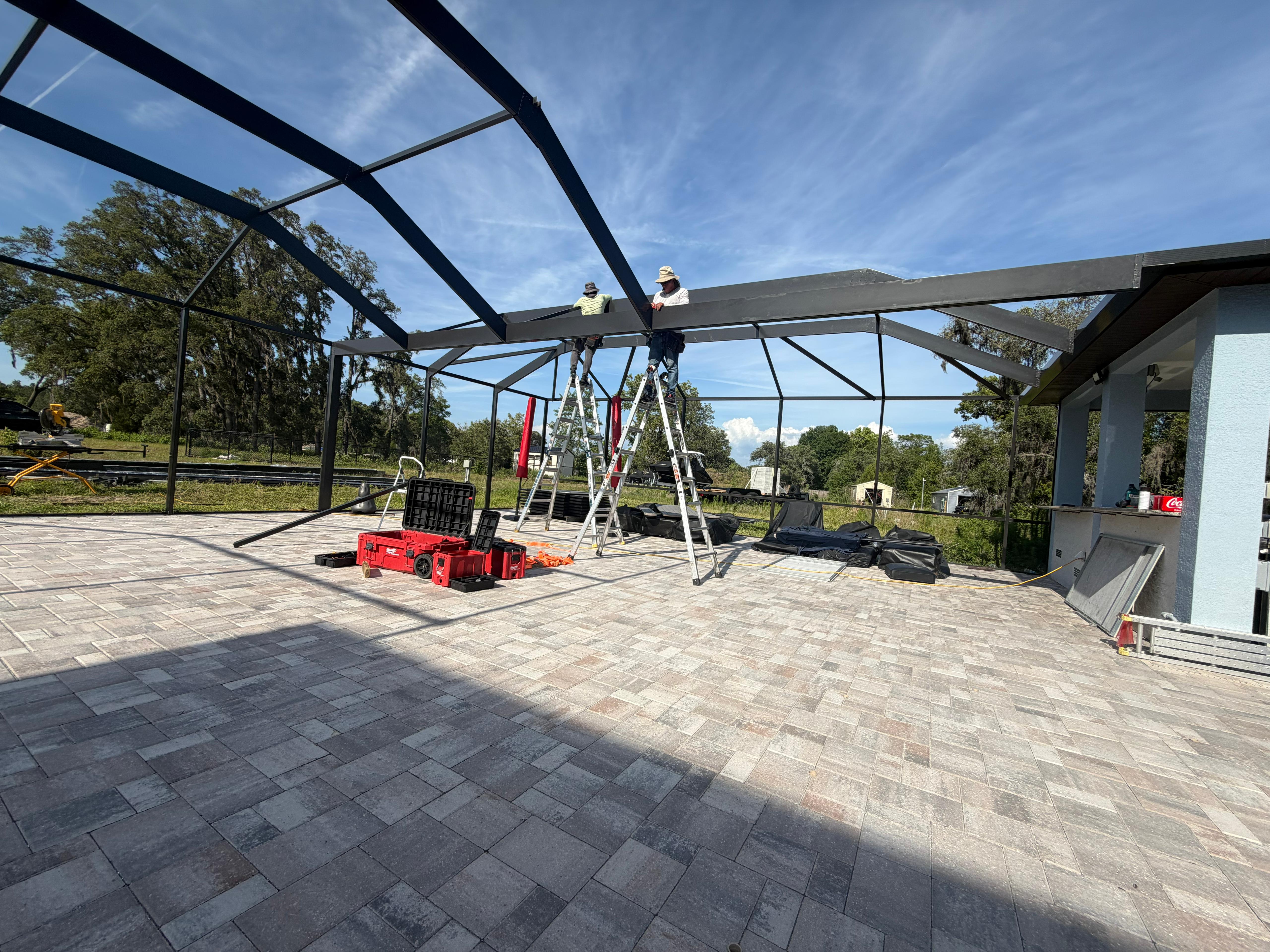 Top Tier Home Building Solutions installation team finishing the mansard roof beams of a custom pool cage in Brooksville, FL — bronze-black powder-coated aluminum, premium build ($80,000–$90,000 tier) (#CGC1527718).