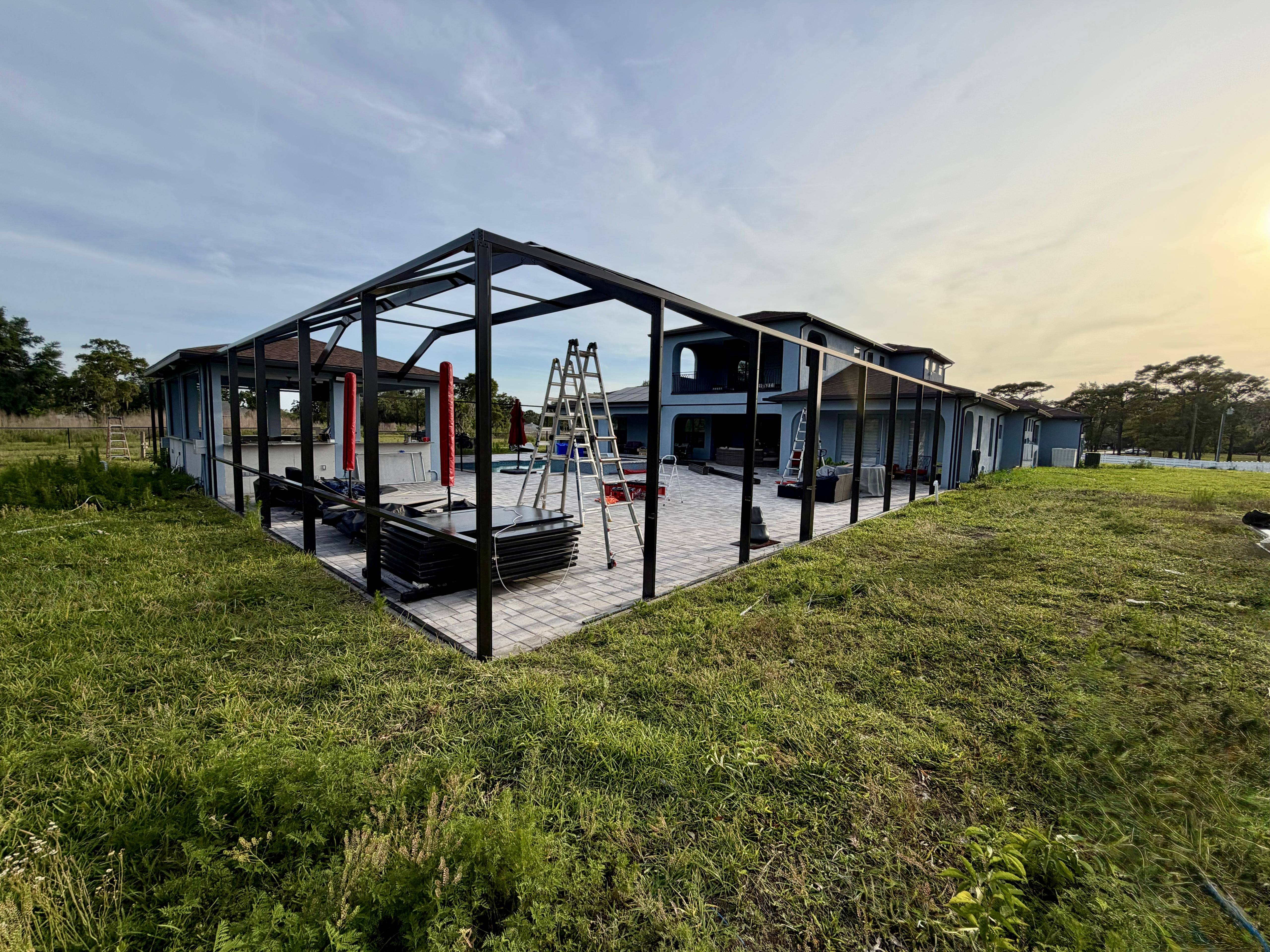 Completed mansard pool cage frame at golden hour in Brooksville, FL — full bronze-black aluminum structure with oversized posts, panoramic side walls, and integration with a two-story Mediterranean home. Built by Top Tier Home Building Solutions (#CGC1527718).