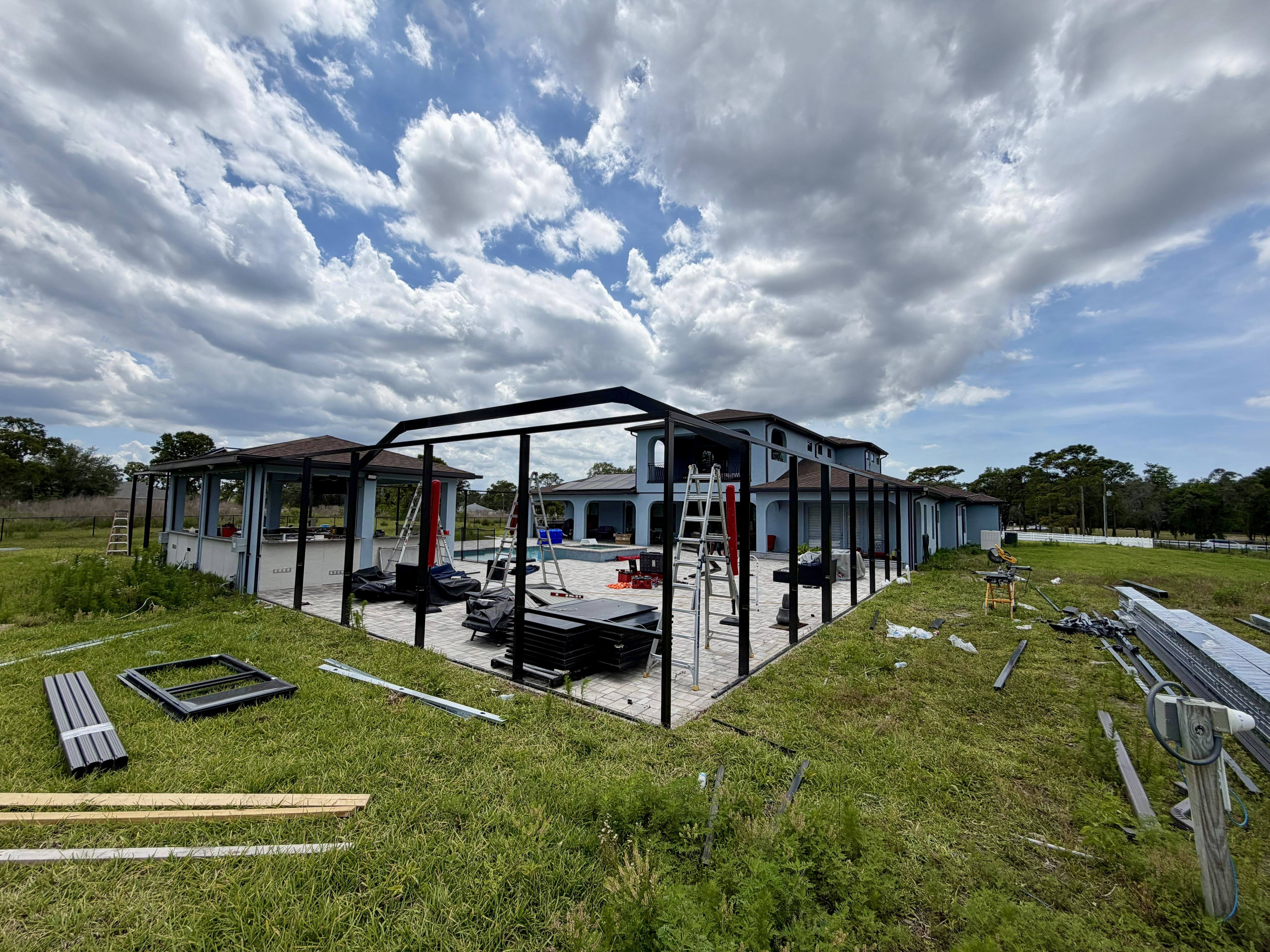Premium 1,100 sq ft mansard pool cage under construction in Brooksville, Florida — bronze-black aluminum framing alongside a two-story Mediterranean home, with existing pool, spa, and covered outdoor kitchen. Built by Top Tier Home Building Solutions (#CGC1527718).