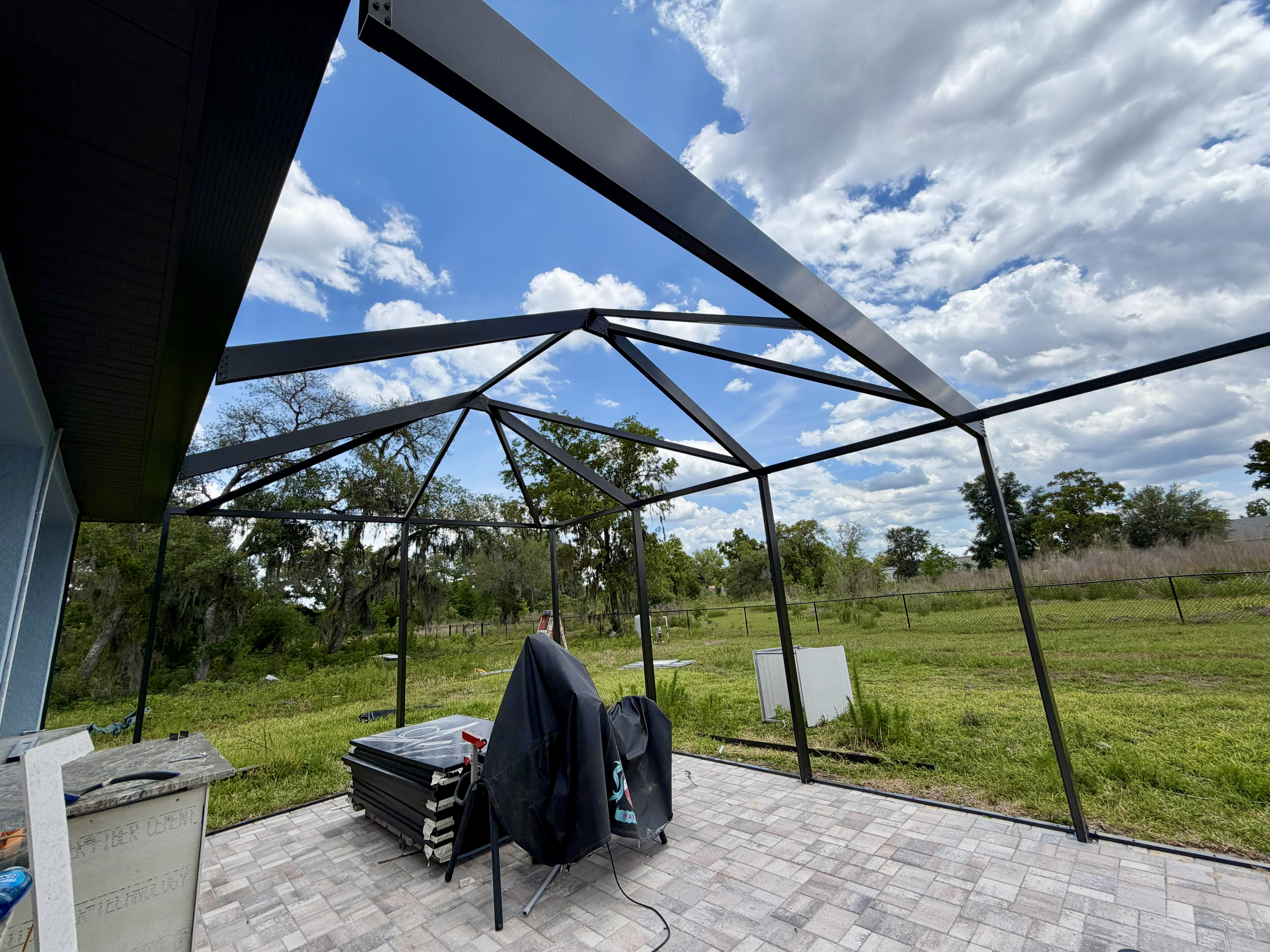 Interior view looking up at the custom mansard roof framing of a premium pool enclosure in Brooksville, FL — bronze-black powder-coated aluminum trusses, hip-roof transition, and oversized structural beams. Top Tier Home Building Solutions (#CGC1527718).